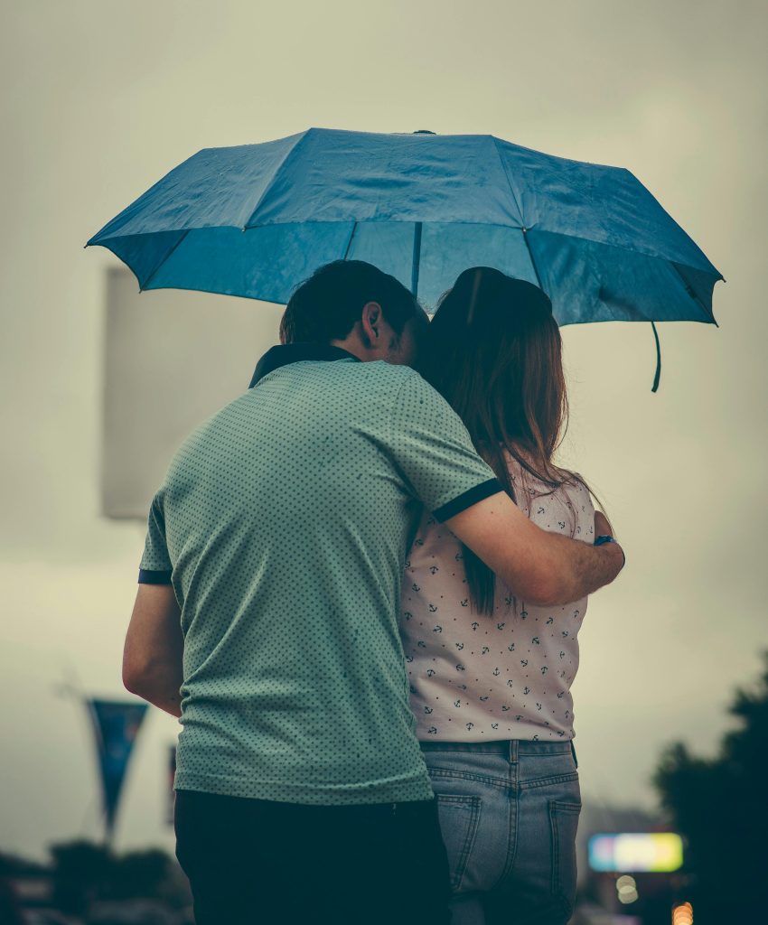pexels-photo-1534633-1534633 A couple embraces under a blue umbrella in a rainy street scene, evoking love and warmth.
